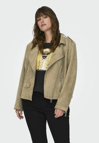 Chaqueta de moto de ante beige con herrajes plateados, bolsillos con cremallera y un diseño corto, llevada sobre una camiseta gráfica negra y jeans oscuros.