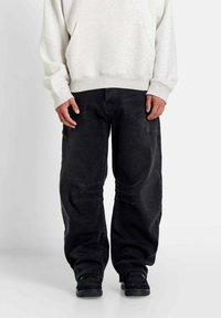 Zwarte cargobroek met een losse pasvorm, voorzien van versterkte knieën en zijzakken. Gemaakt met een lichtgrijze hoodie en zwarte schoenen.
