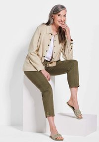 Beige knoopjasje met zwarte knopen, witte top, olijfgroene cropped broek en lichtgrijze slides, zittend op een wit voetstuk.