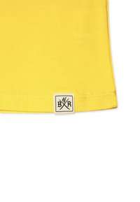 Gul bomulds T-shirt med en syet kant og et lille hvidt mærke med sorte grafikker placeret i nederste venstre hjørne.