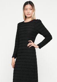 Vestido preto de manga longa com um padrão texturizado e pregas horizontais. O tecido tem um acabamento ligeiramente amarrotado.