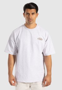 Hombre con cabello oscuro que lleva una camiseta de gran tamaño en un gris claro con un pequeño logo beige en el pecho, de pie contra un fondo sencillo.