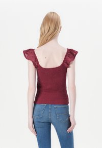 Burgundy top with short ruffled sleeves, smocked texture, and scoop back, paired with blue jeans featuring back pockets and stitching details.