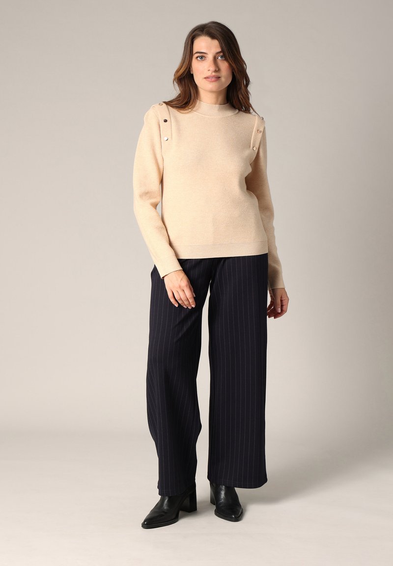 Pull en tricot beige avec texture côtelée et boutons décoratifs sur les épaules. Associé à un pantalon large à rayures bleu marine et des bottes noires à talons.