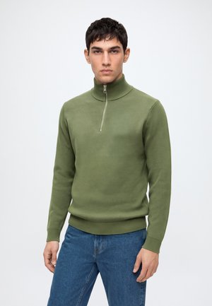 Jeune homme portant un pull vert olive à fermeture éclair sur le devant et un jean bleu, debout devant un fond clair uni, regardant devant lui.
