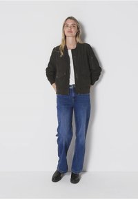 Tmavě olivově zelená bomber bunda s předními kapsami, v kombinaci s světle modrými zvonovými džíny a černými kotníkovými botami. Jednoduchý, neformální styl.