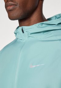 Světlomodrá větrovka s kapucí, s stříbrným logem Nike na přední straně. Hladký povrch s plným zipem.