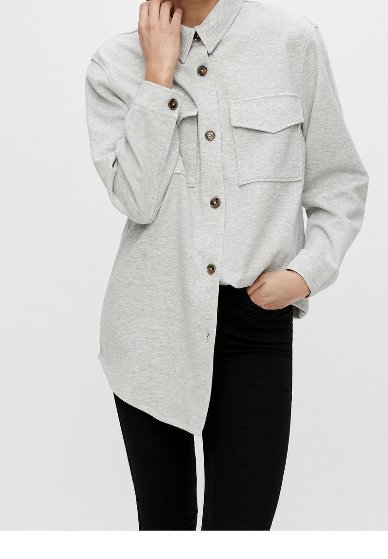 Lys grå skjorte med knapper i blødt stof, to brystlommer, afrundet kant og brune knapper. Matchet med sorte bukser.