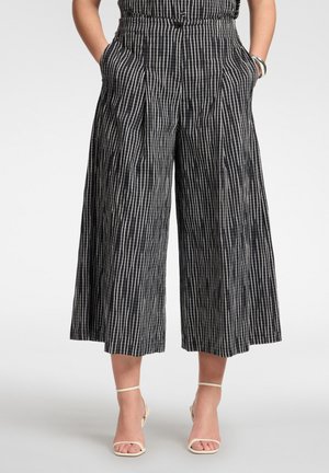 Schwarze, weit geschnittene, verkürzte Hose mit weißen vertikalen Streifen, getragen mit beigen offenen High Heels, Hände in den Taschen und einem elastischen Bund auf Taillenhöhe.