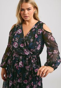 Lång, blommig omlottdress i mörkgrön med lila och blå blommor. Har kalla axlar och ett knyt i midjan. Genomskinlig textur och flödande design.