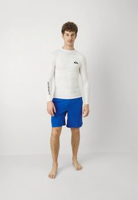 Vitt långärmat rashguard med logotyp på bröstet, kombinerat med blå badshorts. Slätt tyg, figursytt snitt och lätt textur.