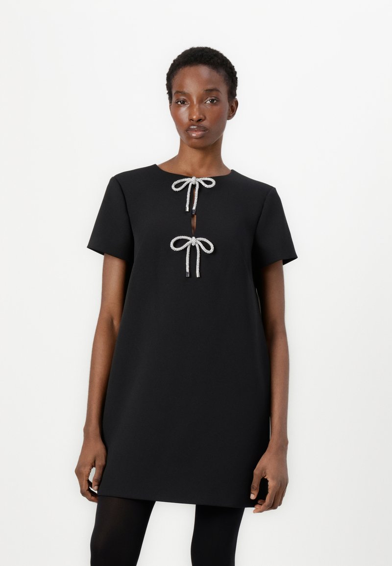 Abito nero a maniche corte con scollatura rotonda, caratterizzato da due fiocchi decorativi bianchi e argentati sul davanti e un delicato taglio nella parte inferiore.