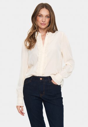 Cremefarvet bluse med flæse ved knappelukningen og manchetterne, letvægtigt stof, parret med mørkeblå jeans med kontrastsyninger og klassisk pasform.