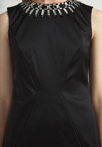 Schwarzes ärmelloses Kleid mit tailliertem Oberteil, das einen mit Perlen verzierten Ausschnitt mit klaren und schwarzen Verzierungen aufweist, glatte Stoffoberfläche.