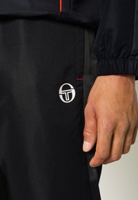 Pantalones deportivos negros en una tela texturizada con un logo plateado y detalles en rojo. Se pueden ver puños elásticos y un detalle de bolsillo lateral.