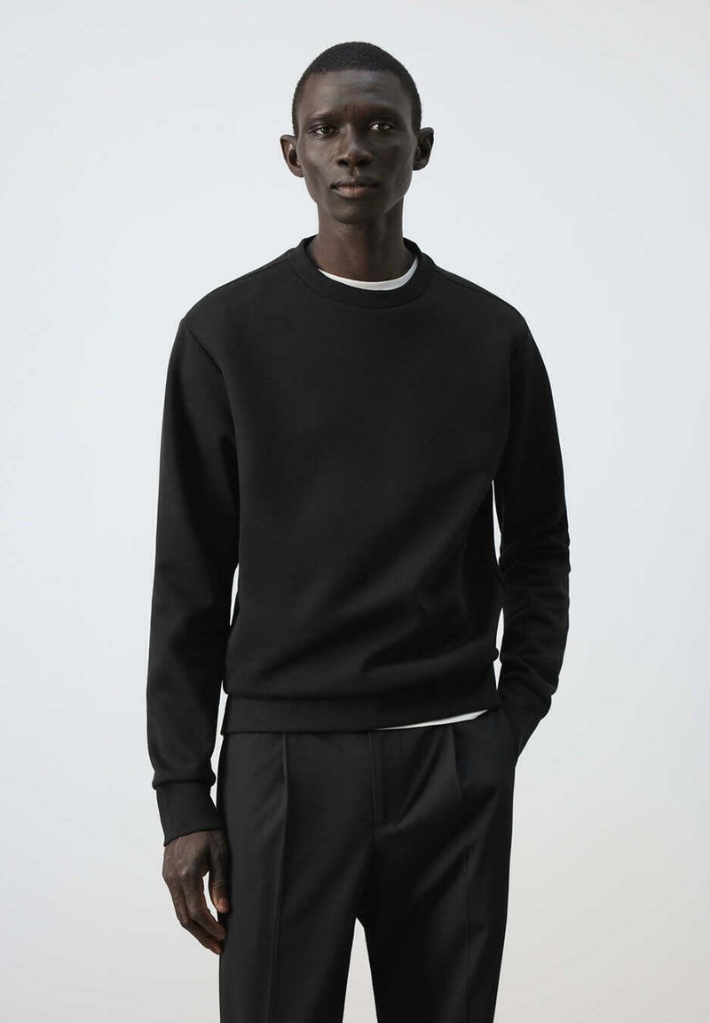 Homme portant un sweat-shirt noir à manches longues et un pantalon noir, debout devant un fond clair uni.