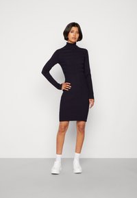 Abito a maglia a coste di colore blu navy con collo alto, maniche lunghe e trame diverse; abbinato a sneakers bianche e calzini alla caviglia.