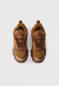 Καφέ αθλητικά παπούτσια με αφράτη υφή, χρυσές λεπτομέρειες και χοντρή σόλα. Περιλαμβάνουν καφέ κορδόνια και στοιχεία branding στη γλώσσα και τη φτέρνα.