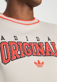 Sudadera gris claro con cuello de canalé rojo. Presenta un llamativo texto en negro y rojo que dice "ADIDAS ORIGINALS" y un pequeño logo adidas rojo.