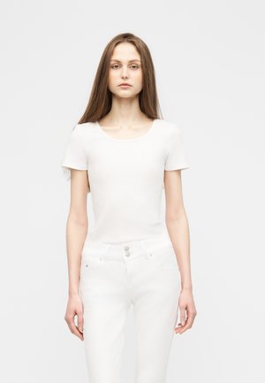 Γυναίκα με μακριά καστανά μαλλιά, φορεμένη με λευκό κοντομάνικο πουκάμισο και λευκό παντελόνι, στέκεται μπροστά σε απλό ανοιχτό γκρι φόντο.