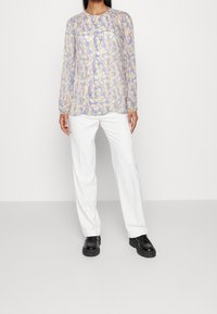 Blouse légère à manches longues avec un motif abstrait jaune, violet et bleu. Portée avec un pantalon blanc et des chaussures noires, elle présente un col rond.