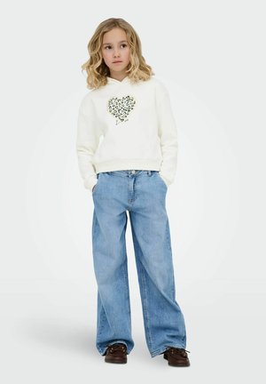 Mergina su šviesiais, pečius siekiančiais plaukais vilki baltą gobtuvinį megztinį su leopardo širdies grafika, plačias mėlynas džinsines kelnes ir rudos spalvos batus, rankos kišenėse.