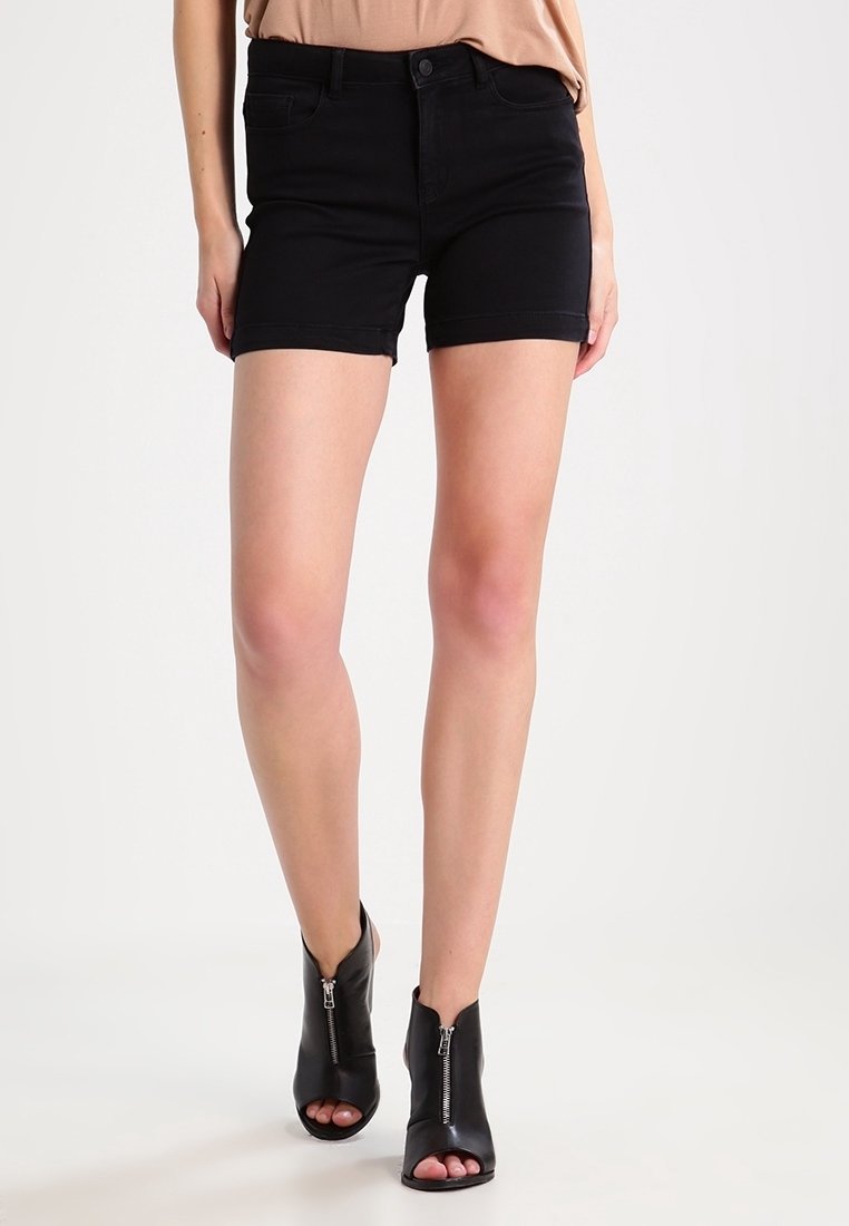 Femme portant un short en jean noir et des bottines noires à bout ouvert avec des fermetures éclair à l'avant, debout devant un fond clair uni.
