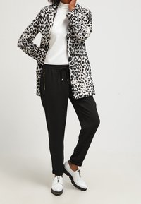 Persona vistiendo un suéter de cuello alto blanco, pantalones negros con cordón, zapatos de plataforma blancos y un abrigo de estampado de leopardo en blanco y negro, de pie con la mano cerca de la cara.
