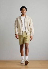 Μπεζ σακάκι με ελαστικά μανσέτα, λευκό μπλουζάκι, κίτρινα σορτς με υφασματικό σχέδιο και ανοιχτά αθλητικά παπούτσια με ρίγες και λευκές κάλτσες.