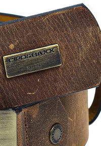 Brown leather bag with a textured surface, featuring a gold-tone metal tag with "Gipo&BaXX" and "Est. 1996," rounded edges, and a circular button detail.