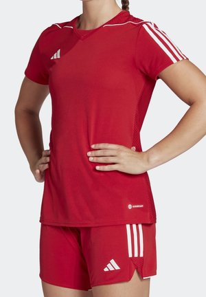 Femme portant un maillot et un short de sport rouges Adidas avec des bandes blanches, debout les mains sur les hanches sur un fond uni.