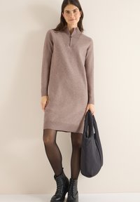 Světle taupe pletené šaty s vysokým límcem a zipem, ribkované manžety a lem. Model drží černou volnou kabelku a má na sobě černé kotníkové boty.