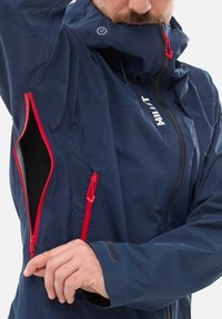 Granatowa wodoodporna kurtka z czerwonymi zamkami, regulowanym kapturem i kieszeniami po bokach; ma teksturowany materiał oraz mankiety na rzep.