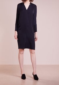 Tummansininen, polvipituinen mekko, jossa on V-pääntie, pitkät hihat ja napitusyksityiskohta kauluksessa. Valmistettu pehmeästä, kevyestä materiaalista.