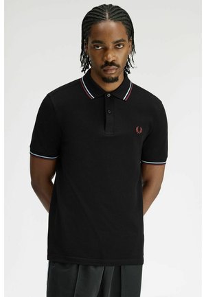 Uomo con capelli intrecciati che indossa una polo nera con colletto e polsini a righe rosse e blu, in piedi con le mani dietro la schiena.