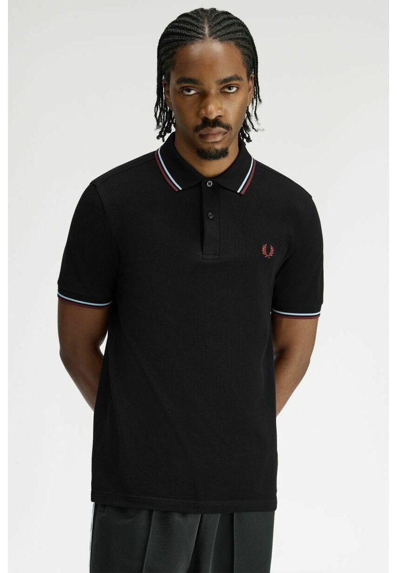 Homme aux cheveux tressés portant un polo noir avec un col et des poignets de manches rayés rouge et bleu, debout les mains derrière le dos.