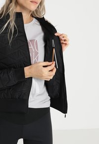 Svart kviltad jacka med en ficka med dragkedja som har en orange dragrem, bärs över en ljusgrå t-shirt. Slätt tyg, figursydd design.