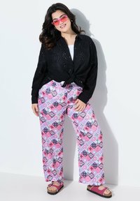Schwarzes Augenlochkastenhemd mit geknoteter Vorderseite, kombiniert mit weit geschnittenen, rosa gemusterten Hosen mit geometrischen Designs. Rosa Sonnenbrille und schwarze Plateausandalen.