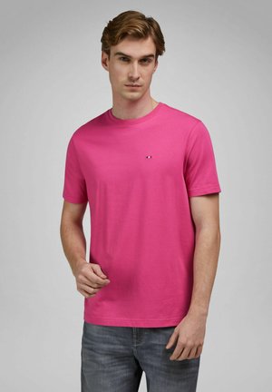 Jonge man met lichtbruin haar, gekleed in een felroze T-shirt met korte mouwen en een blauwe spijkerbroek, tegen een effen grijze achtergrond.