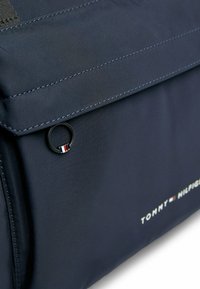 Borsa in nylon blu navy con chiusura a pattina, caratterizzata da linee cucite orizzontali e un piccolo logo circolare con accenti rossi, bianchi e blu.