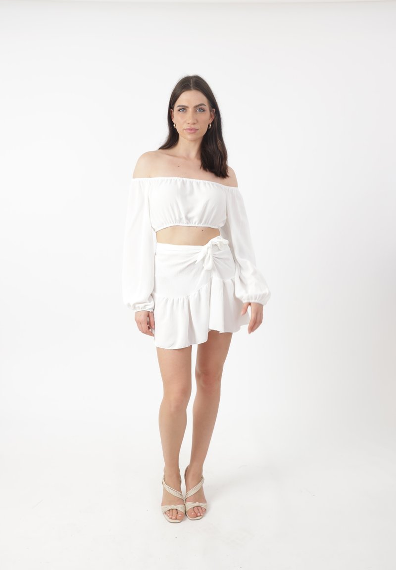 Weißes schulterfreies Crop-Top mit puffigen Ärmeln, kombiniert mit einem weißen Minirock mit Bindeband und ausgestelltem Saum. Neutrale Riemchensandalen.