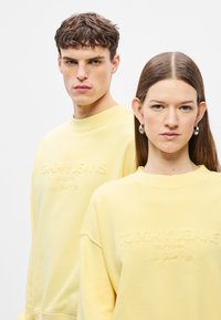 Κίτρινο φούτερ με ανάγλυφο λογότυπο "TOMMY JEANS", στρογγυλή λαιμόκοψη και άνετη εφαρμογή, φορεμένο από δύο μοντέλα σε ουδέτερο φόντο.