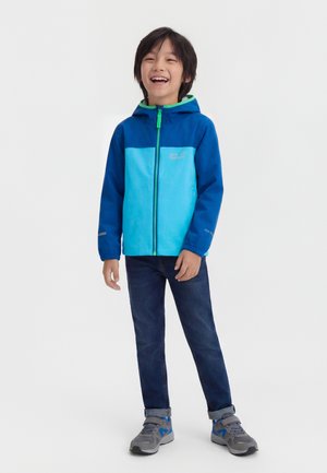 Enfant souriant portant une veste à capuche bicolore bleue, un jean foncé et des baskets grises, debout devant un fond clair uni.