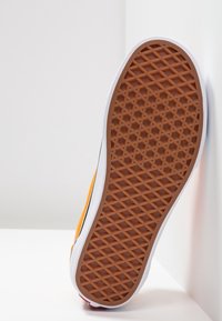 Semelle de chaussure avec une surface en caoutchouc marron présentant un motif hexagonal texturé. Côté blanc, section supérieure orange et accents rouges visibles.