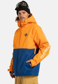 Mężczyzna w czarnej czapce beanie i goglach narciarskich, w pomarańczowej i niebieskiej kurtce z logo oraz beżowych spodniach, stojący z jedną ręką w kieszeni.