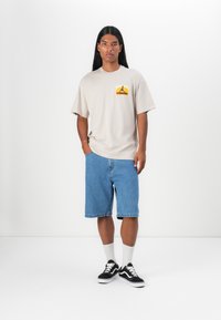 Beige oversized t-skjorte med et fargerikt grafisk motiv på venstre bryst, sammen med lyseblå denimshorts og svarte sneakers med hvite detaljer.