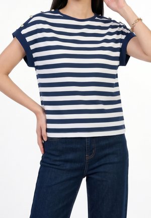 Vrouw draagt een navyblauw-wit horizontaal gestreept kort mouw shirt met knoopdetails op de schouders en donkerblauwe jeans.