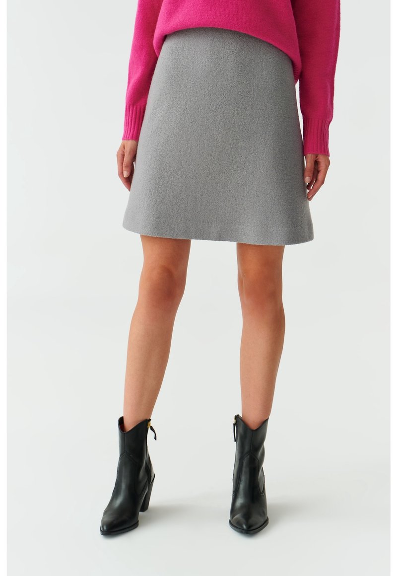 Femme portant une jupe grise au genou, un pull rose et des bottines noires, debout sur un fond clair.