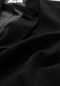 Svart skräddarsydd kavaj med synlig slag och slät inre foder, visad i en närbild från en sned vinkel.