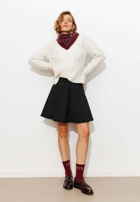 Pull en tricot blanc avec un motif en cablage, jupe noire évasée, écharpe bordeaux, et chaussettes assorties avec le texte "1975", accompagnées de chaussures sombres.